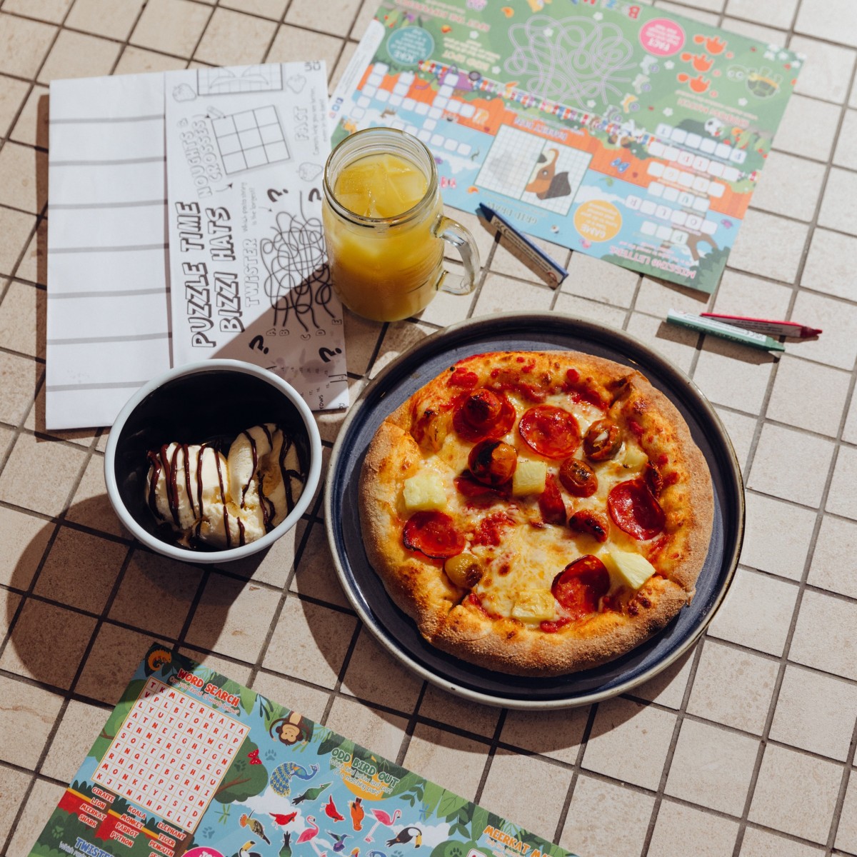 A pepperoni and pineapple pizza, a bowl of ice cream and a glass of orange juice on a table with kids puzzles